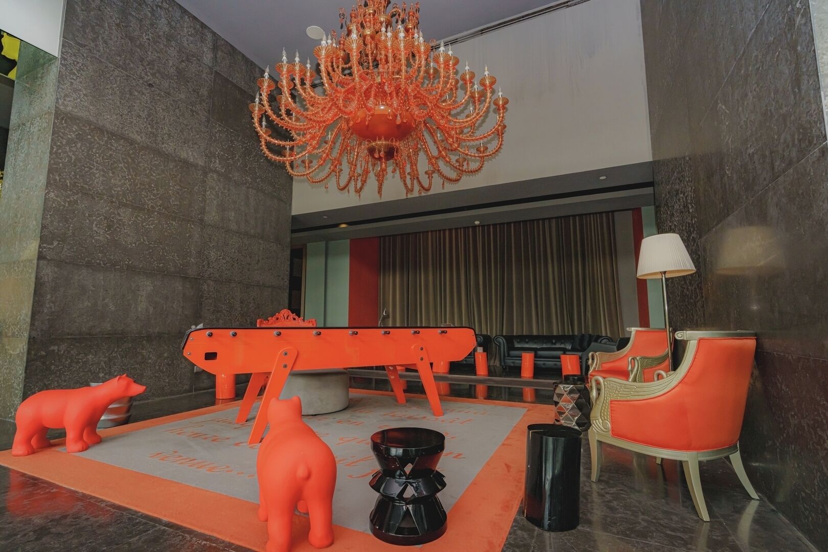 A modern lobby with a large orange chandelier, a seating area with a sofa and armchairs, and a decorative orange sculpture.