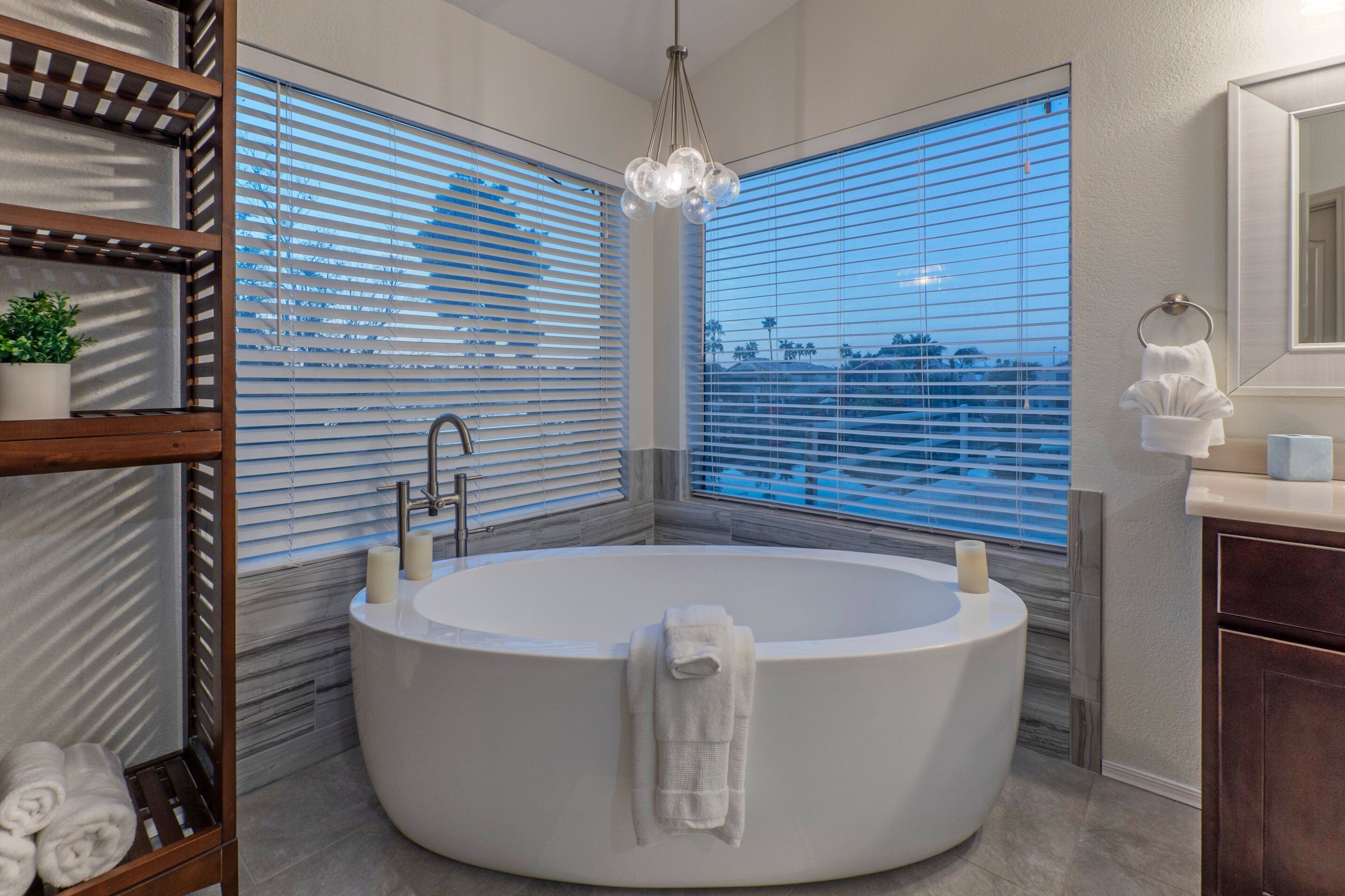Ein modernes Badezimmer mit einer freistehenden Badewanne, Holzregalen mit Handtüchern und einem großen Fenster mit Jalousien.