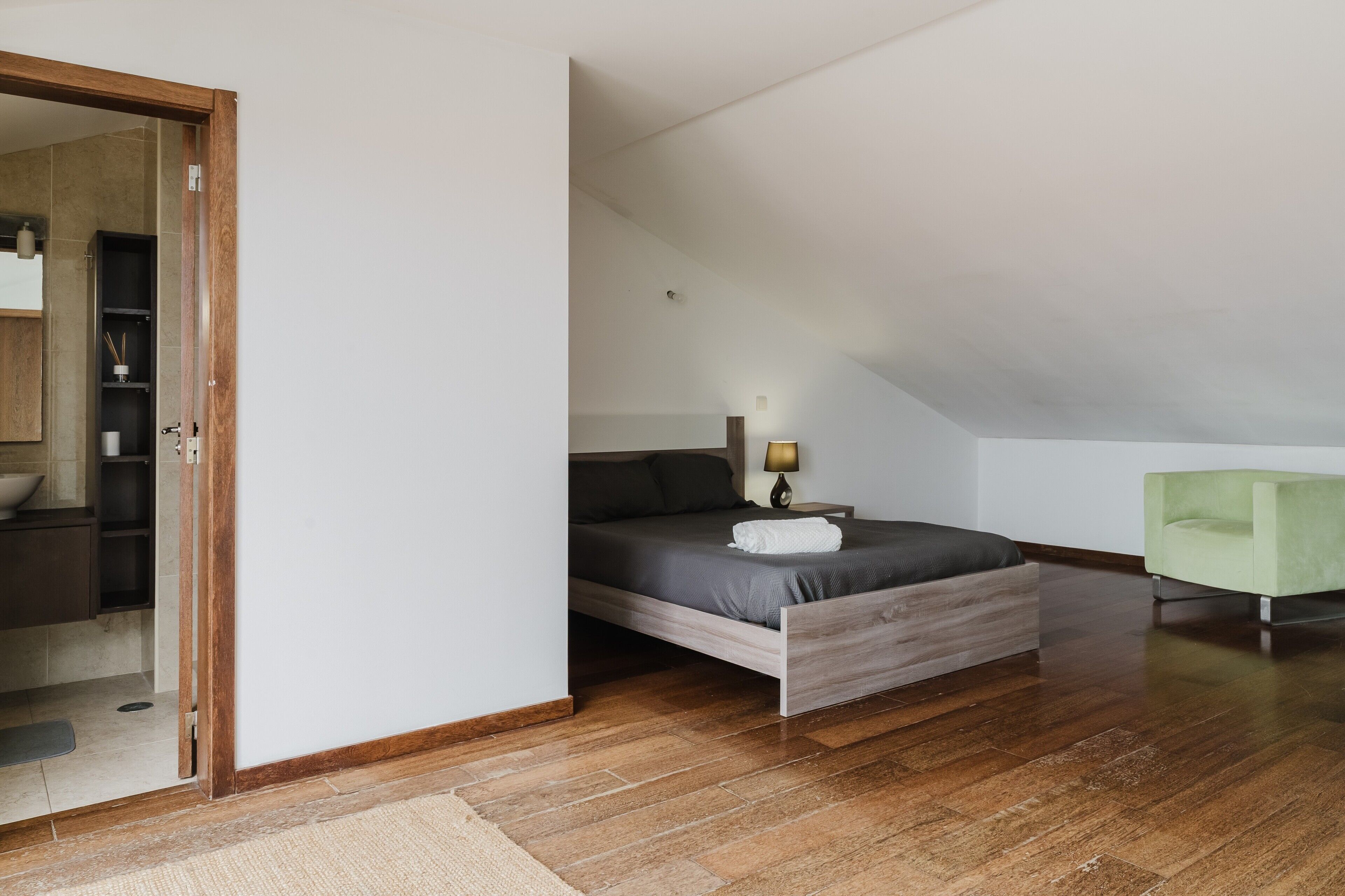 Une chambre à coucher avec un lit en bois, une chaise verte et une salle de bain visible, équipée d’un lavabo et d’un miroir.