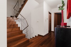 Una escalera de madera con barandales metálicos que conduce a una plataforma, una pared blanca y un florero rojo con flores sobre una mesa negra.