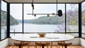 Ein Essbereich mit großem Fenster, das auf einen See und Hügel blickt, inklusive rundem Tisch mit Stühlen und Pendelleuchten.