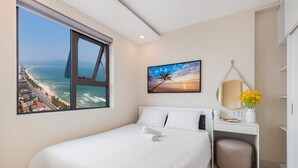 Une chambre d’hôtel dotée d’une grande fenêtre donnant sur la plage et l’océan, d’un lit soigneusement fait, d’un bureau avec un miroir et d’un vase rempli de fleurs.