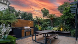 Eine Terrasse im Freien mit einem Holztisch und Bänken, umgeben von üppiger Bepflanzung und einem Grill.
