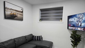 Un salon moderne avec un canapé d’angle en cuir, un grand téléviseur à écran plat, un tableau représentant un paysage encadré, une fenêtre avec des stores et une plante en pot.