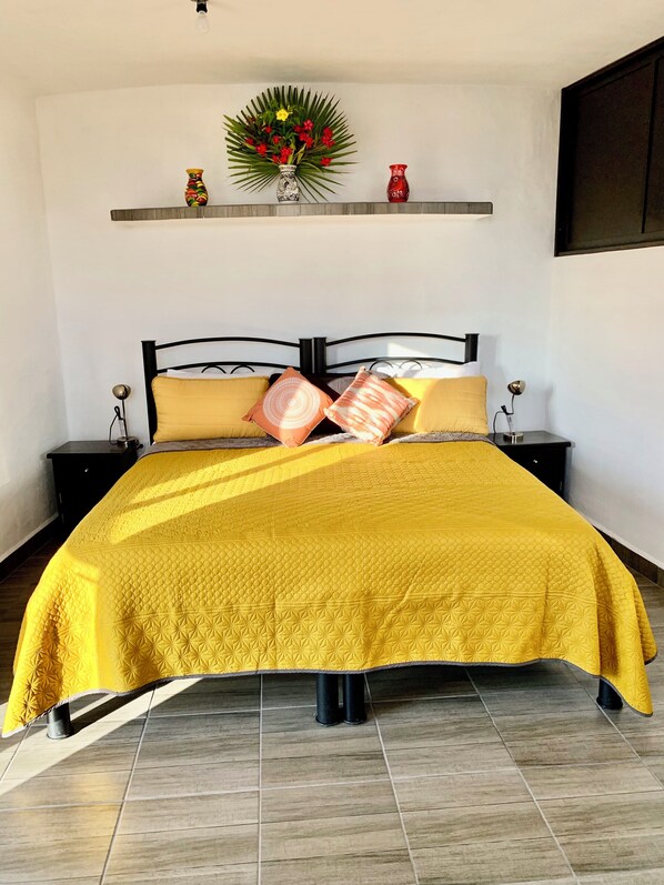 A bed with a yellow quilt, two nightstands, a wall-mounted shelf with decorative items, and a potted plant.