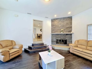 A living room with two leather sofas, a white coffee table with a flower, a stone fireplace, and a staircase leading to another room.