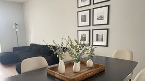 Un coin repas avec une table sombre, des chaises blanches et trois vases blancs contenant des plantes, le tout posé sur un plateau en bois.