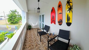 Uma varanda com rede, duas cadeiras, uma mesa pequena e pranchas de surf na parede.