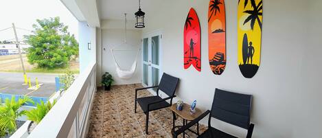 Un balcon avec un hamac, deux chaises, une petite table et des planches de surf accrochées au mur.
