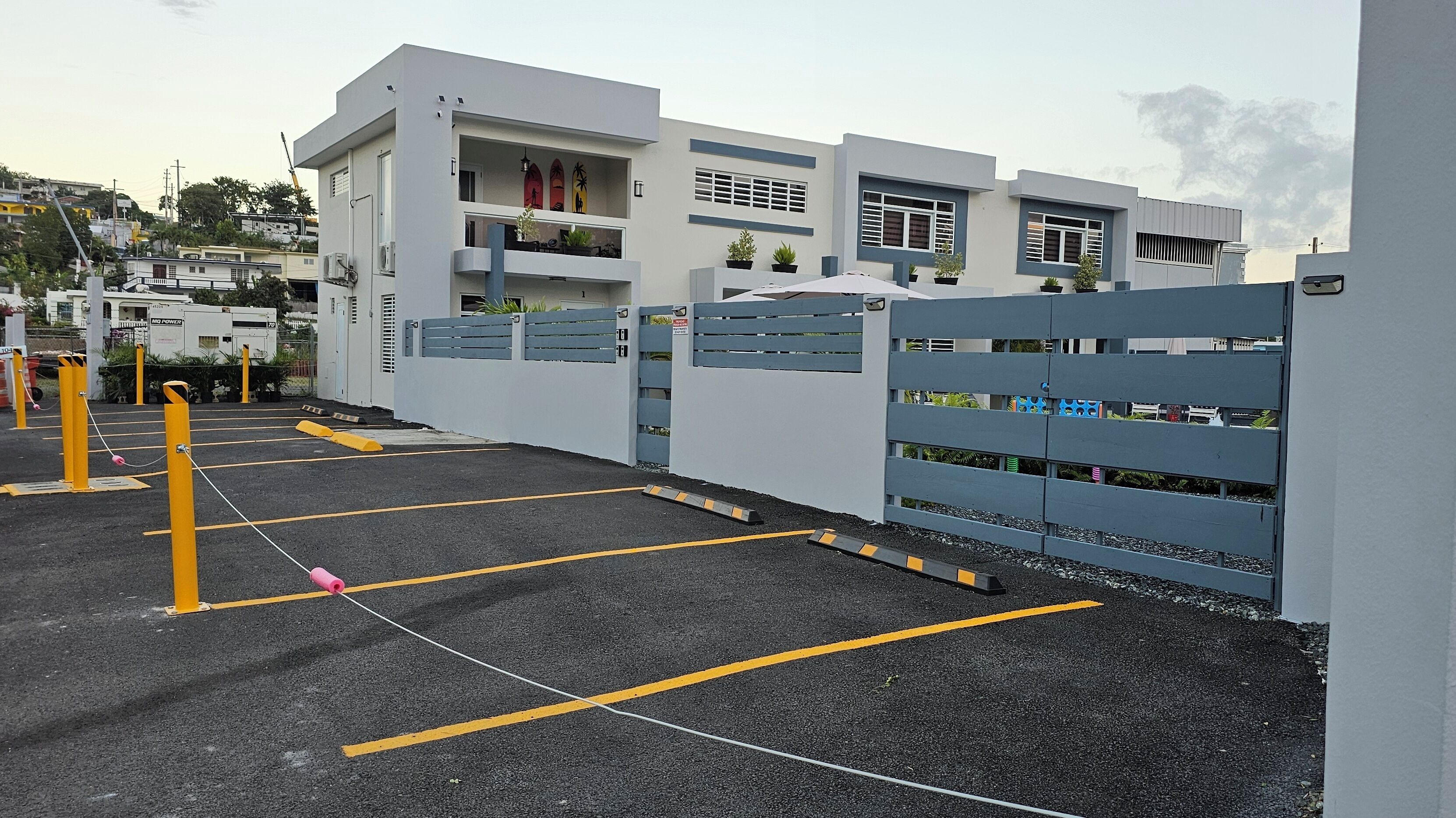 A parking lot with yellow bollards and a gated entrance to a building with balconies and potted plants.