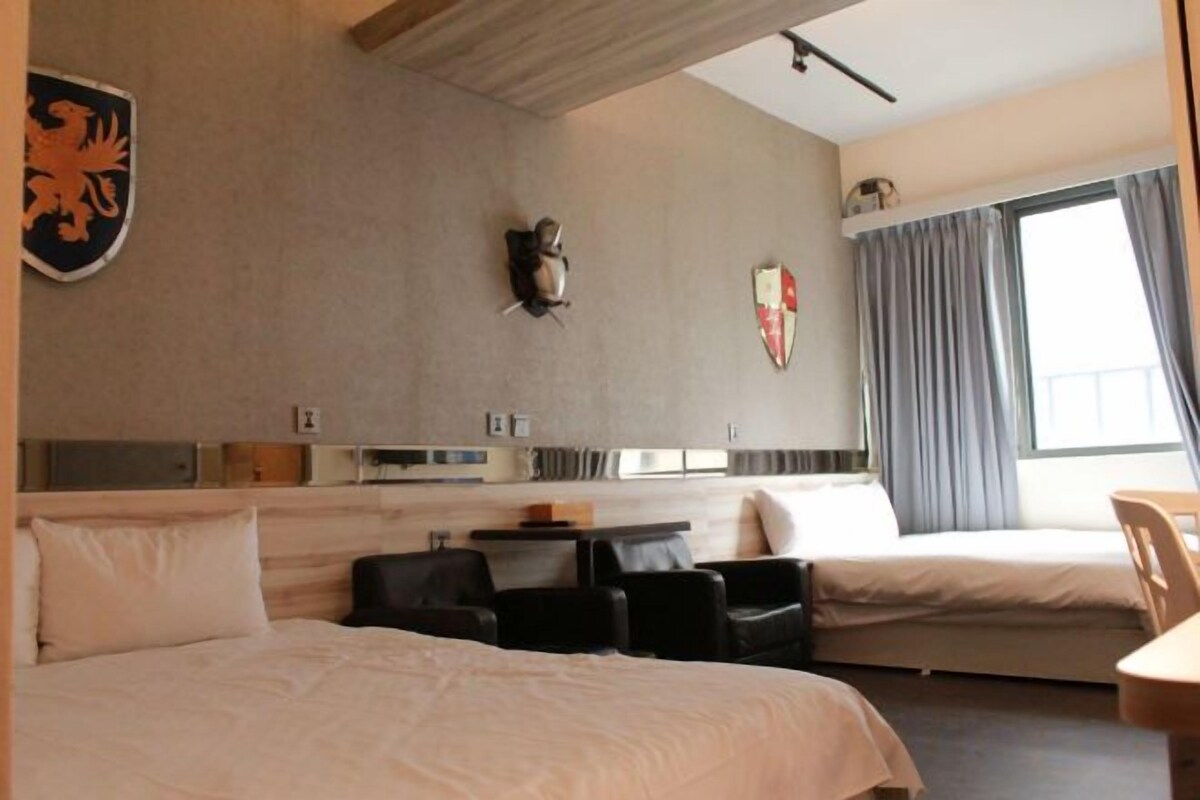 Quarto de hotel com cama, duas cadeiras, uma mesinha e janela com cortinas.