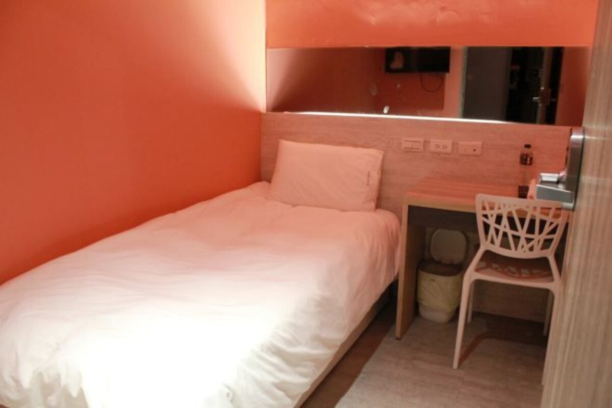 Une petite chambre avec un lit simple, une tête de lit en bois, une table de chevet, une chaise et une poubelle.