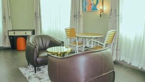 A living room with a brown leather sofa, a small round table, and two chairs. There is a white cabinet and an orange trash bin in the background. - Ahaka cottages  (Mbarara)