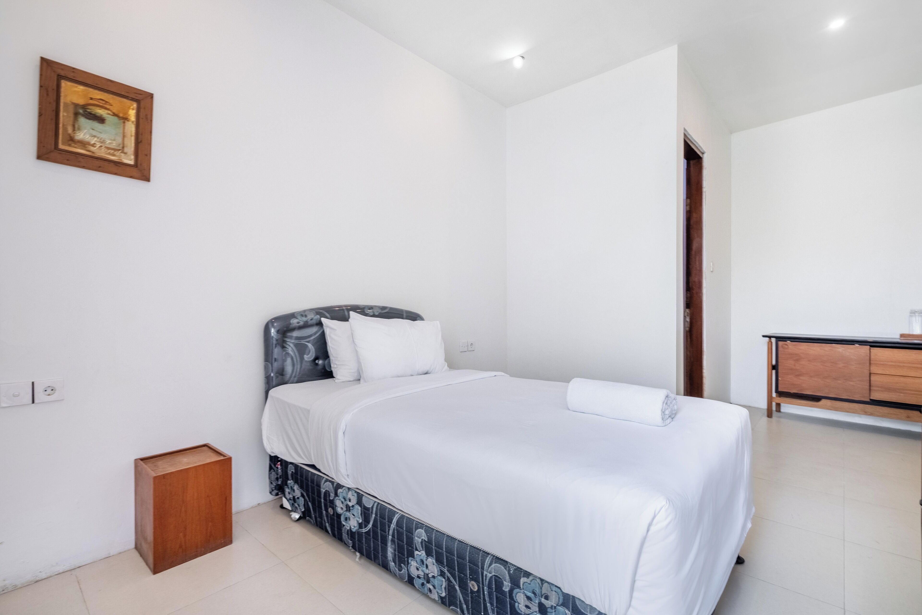 Une chambre à coucher avec un lit, une table de chevet en bois, une commode et un tableau encadré au mur.