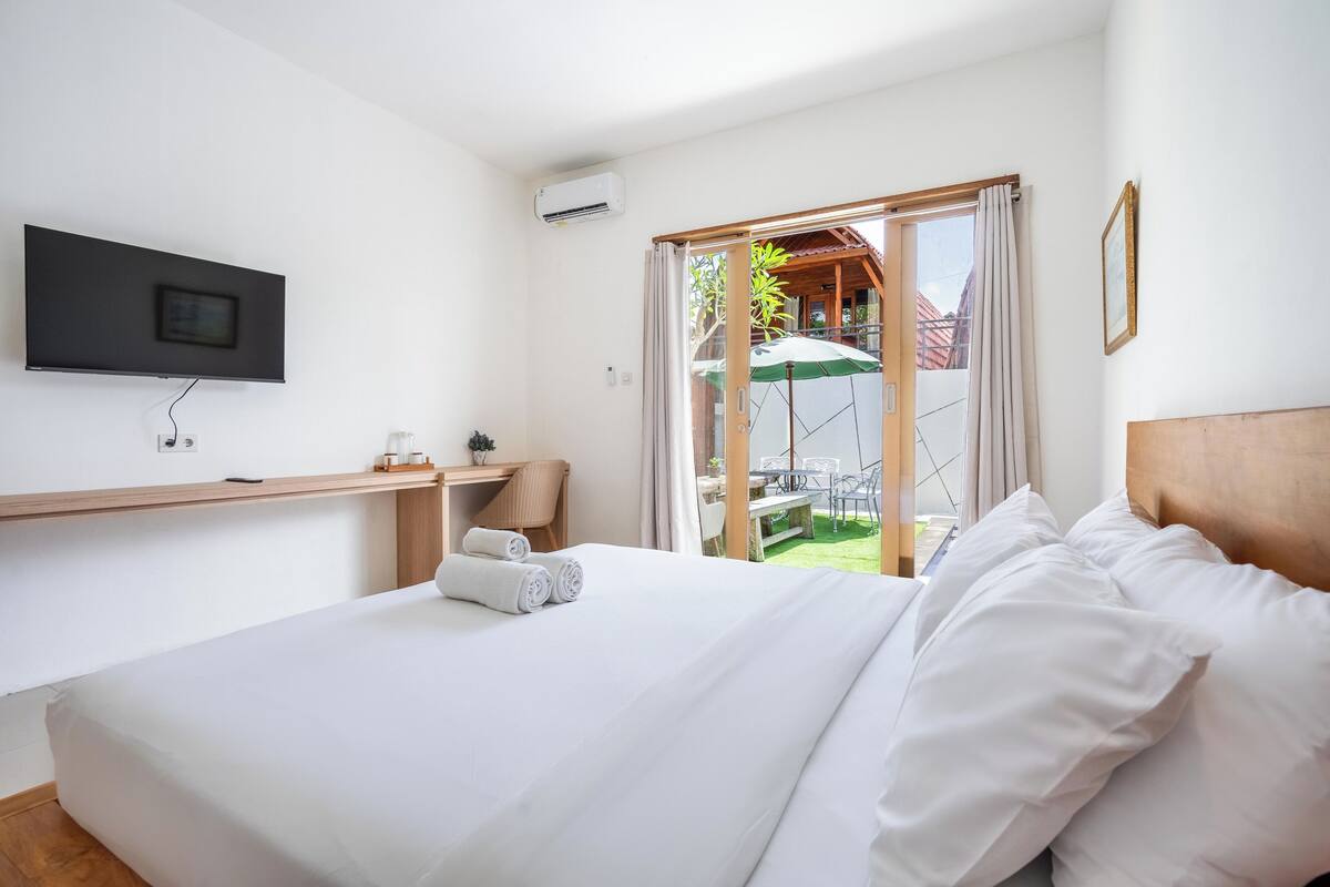 Habitación de hotel con cama, escritorio con silla, televisor en la pared y vista a un área exterior con mesa y sombrilla.