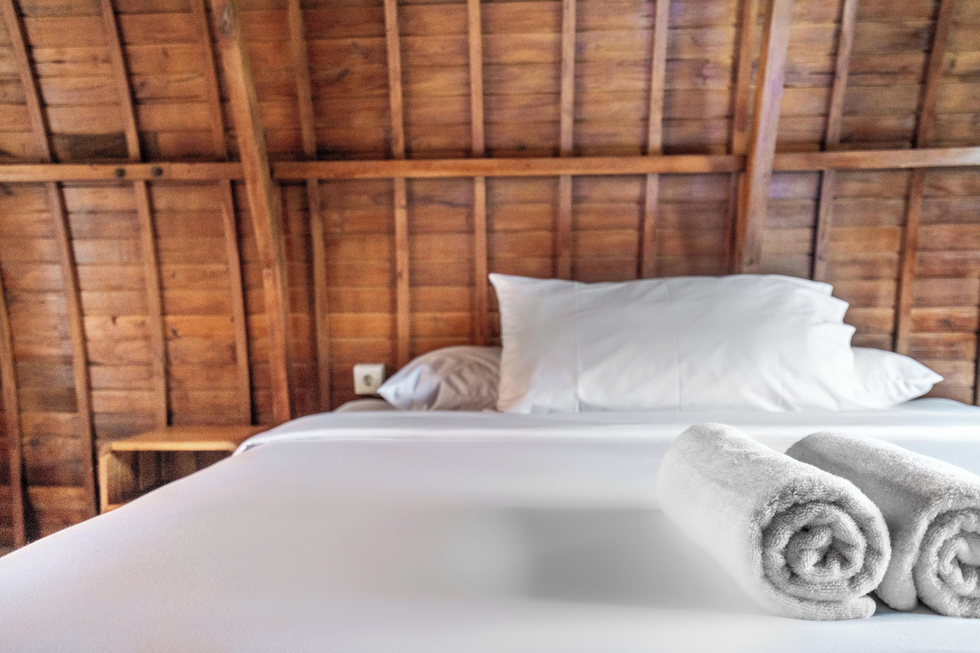 Un lit avec du linge de lit blanc et des serviettes pliées, des poutres en bois au plafond et un petit tabouret en bois à côté du lit.