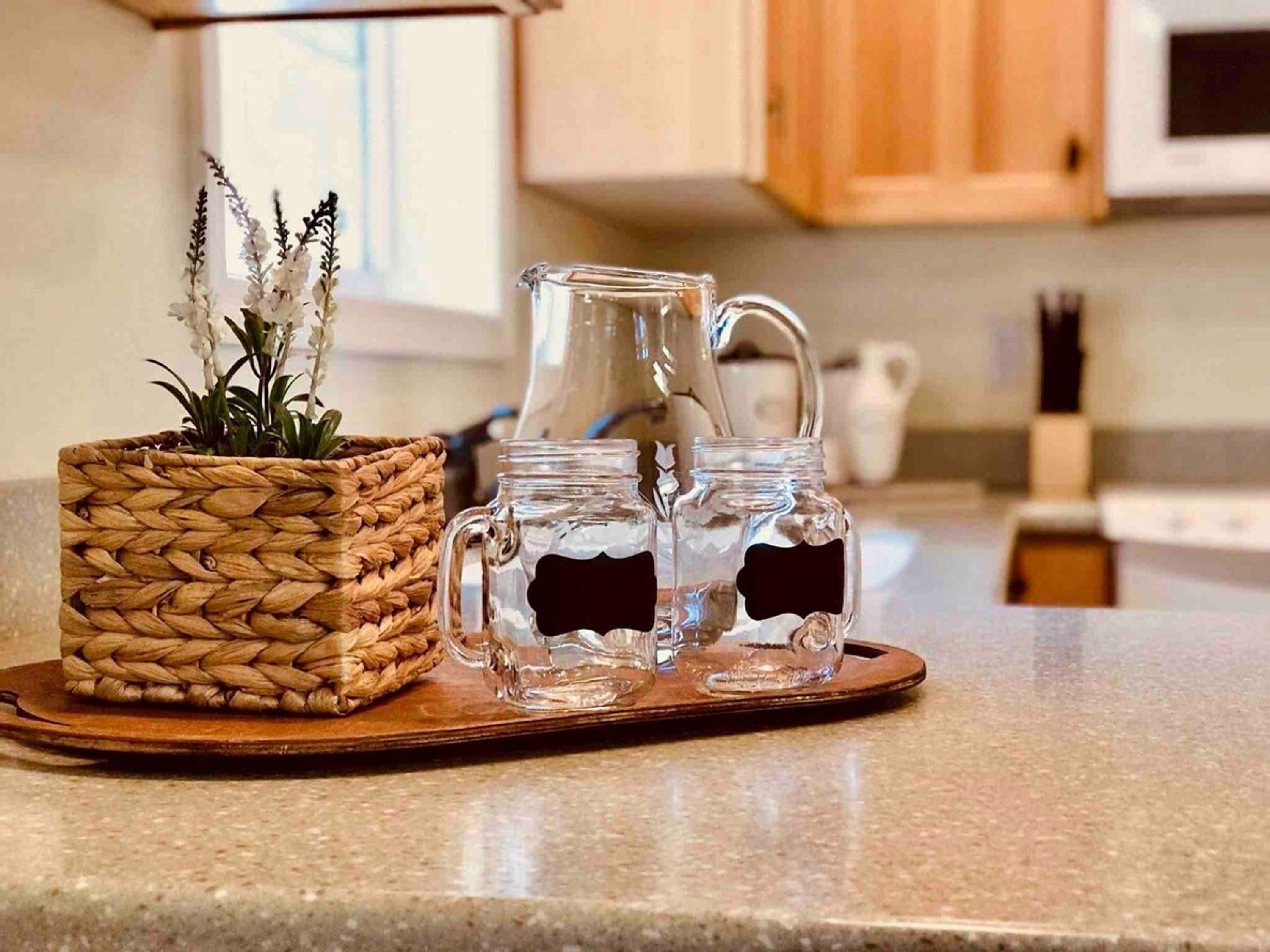 Uma bancada de cozinha com uma cesta de fibra natural contendo flores brancas e dois jarros de vidro com líquido escuro.