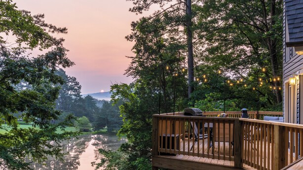 A wooden deck with a view of a calm river and trees, a grill, and string lights.