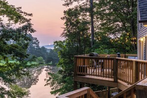 A wooden deck with a view of a calm river and trees, a grill, and string lights. - Lake House near Lookout Mt, Theater, Gym & Arcade (Trenton)