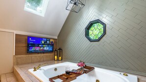 A modern bathroom with a large bathtub, a wooden tray, a glass of wine, and a flower arrangement. A television is mounted on the wall, and there is a window with a hexagonal frame.