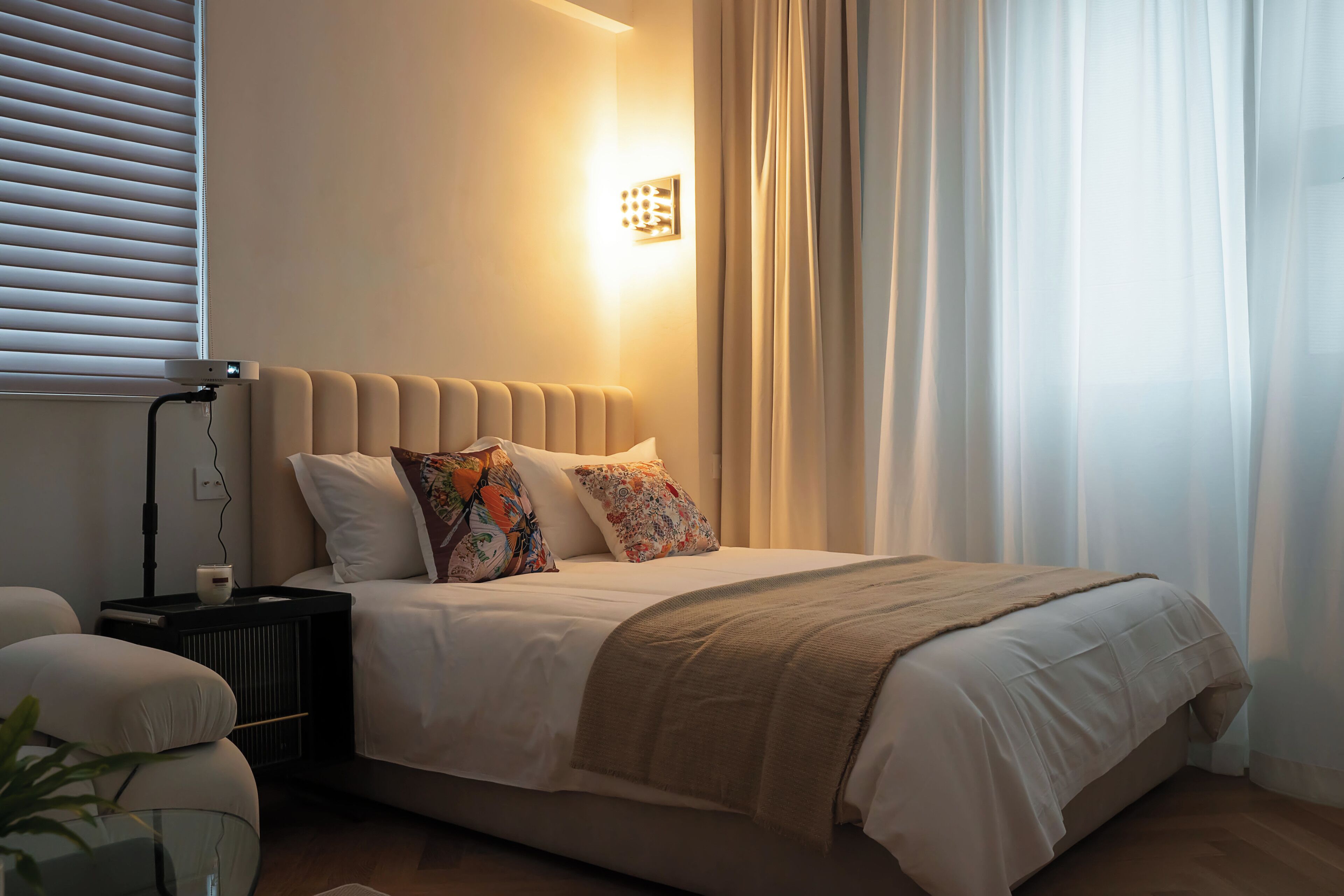 A neatly made bed with a beige headboard, white bedding, and a brown throw blanket. There is a bedside table with a lamp and a small white object. The room has a window with blinds, a curtain, and a wall-mounted light fixture.