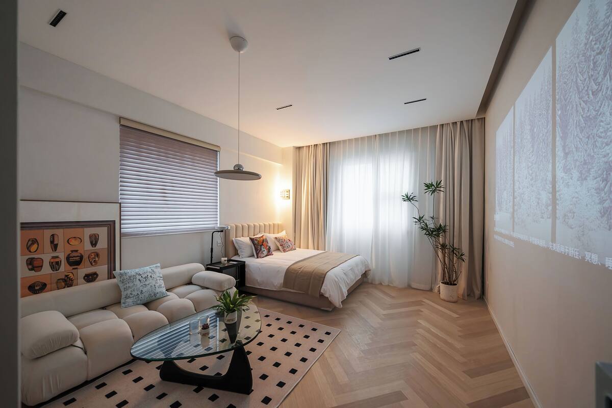 Ein modernes Hotelzimmer mit einer Couch, einem Glastisch, einem Bett mit beiger Decke und einer Zimmerpflanze.
