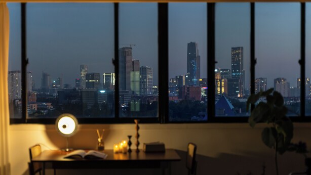 Habitación con escritorio, silla, lámpara y vistas a un horizonte urbano a través de amplios ventanales.