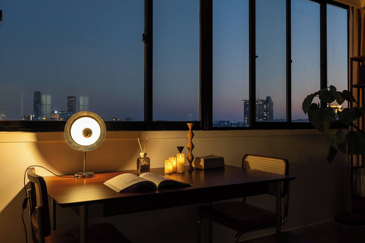 Ein Schreibtisch mit Lampe, Kerzen und einem offenen Buch an einem Fenster mit Blick auf die Stadt.