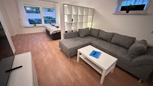 Una sala moderna con un sofá seccional gris, una mesa de centro blanca y una cama con cabecera blanca. Se ve una ventana con cortinas y vistas a árboles fuera.