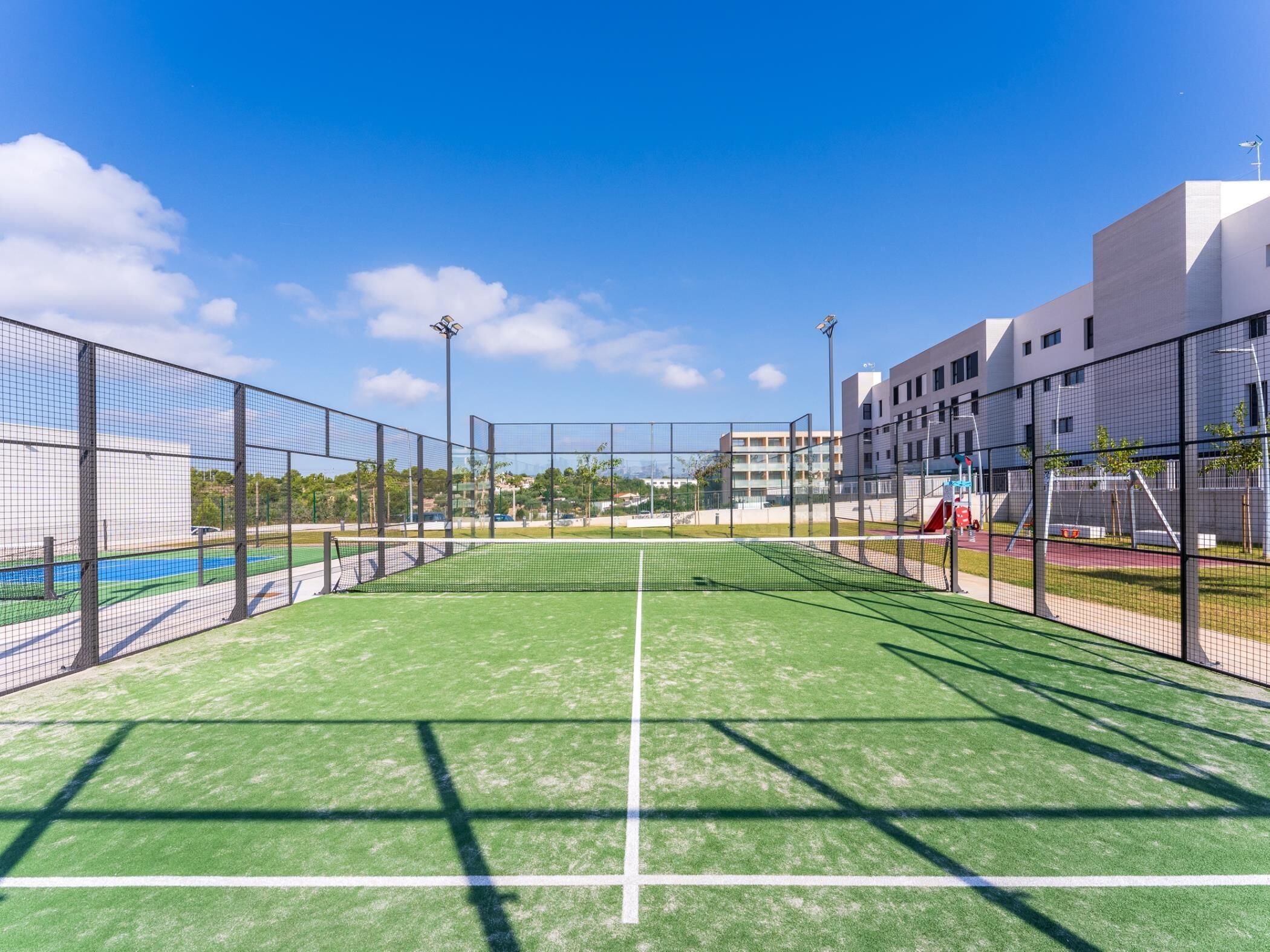 Un court de tennis avec un filet, entouré d’une clôture, et des bâtiments en arrière-plan.
