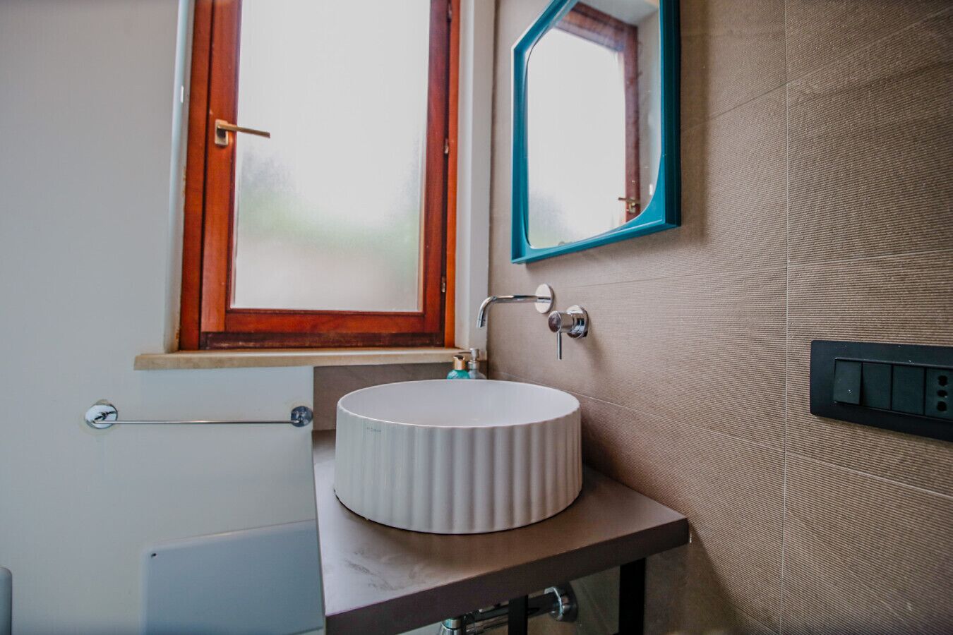 Un baño con un lavamanos redondo, una ventana con marco de madera y un espejo con marco verde azulado.