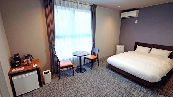 A hotel room with a bed, a desk with a coffee maker, a chair, a small table, and a window with curtains.
