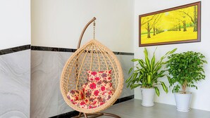 Una silla colgante de mimbre con cojines estampados, una planta en maceta y un cuadro en la pared.