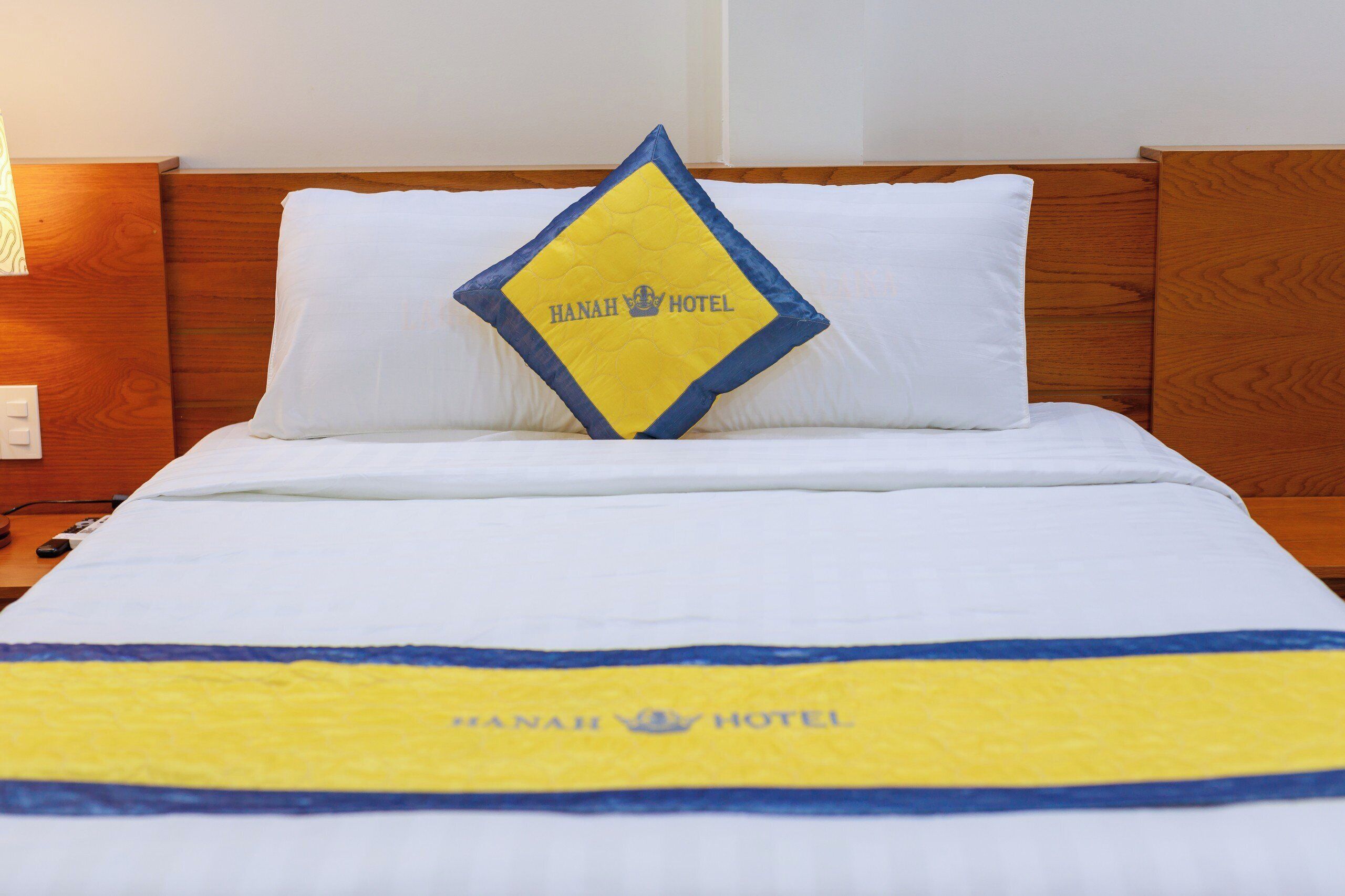 Una cama de hotel con una colcha blanca y amarilla, un almohadón con el logotipo del hotel y un cabecero de madera.
