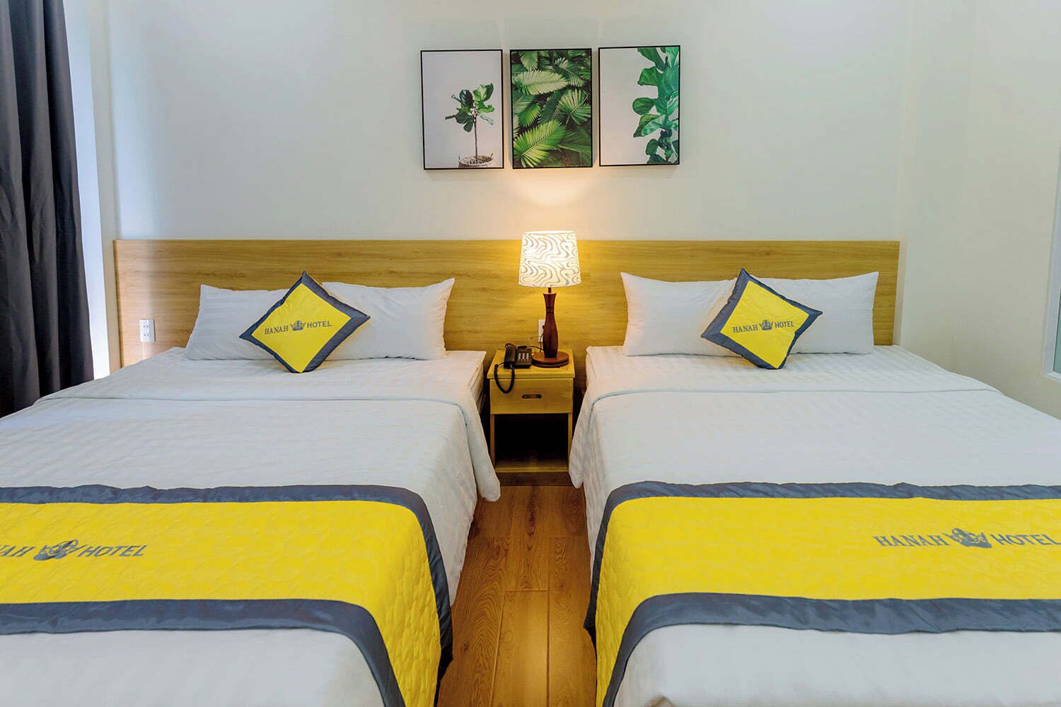 Ein Hotelzimmer mit zwei Betten, jedes mit einer gelb-blauen Tagesdecke und passenden Kissen. Zwischen den Betten befindet sich ein Nachttisch mit einer Lampe. Das Zimmer hat ein Holz-Kopfteil und eine Wand mit drei gerahmten Bildern.