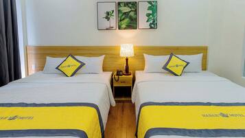 Ein Hotelzimmer mit zwei Betten, jedes mit einer gelb-blauen Tagesdecke und passenden Kissen. Zwischen den Betten befindet sich ein Nachttisch mit einer Lampe. Das Zimmer hat ein Holz-Kopfteil und eine Wand mit drei gerahmten Bildern.