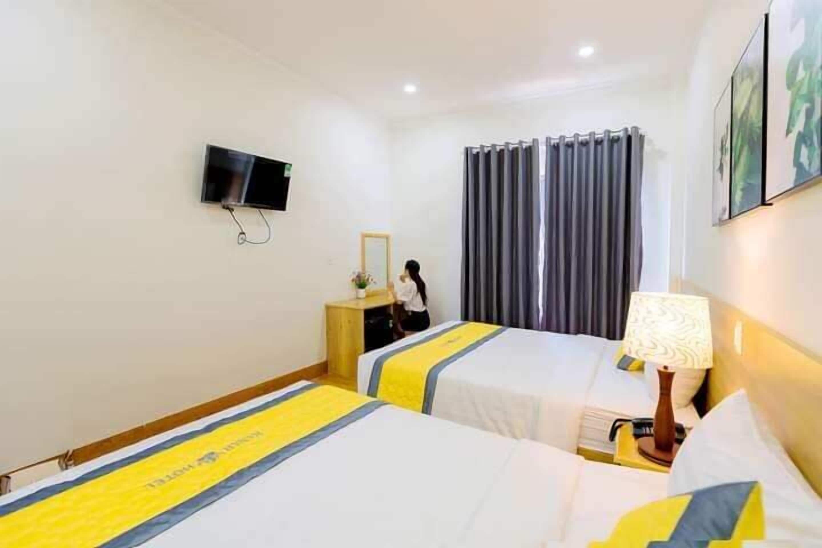 A hotel room with two beds, a TV mounted on the wall, a desk with a mirror, and a lamp on the nightstand.