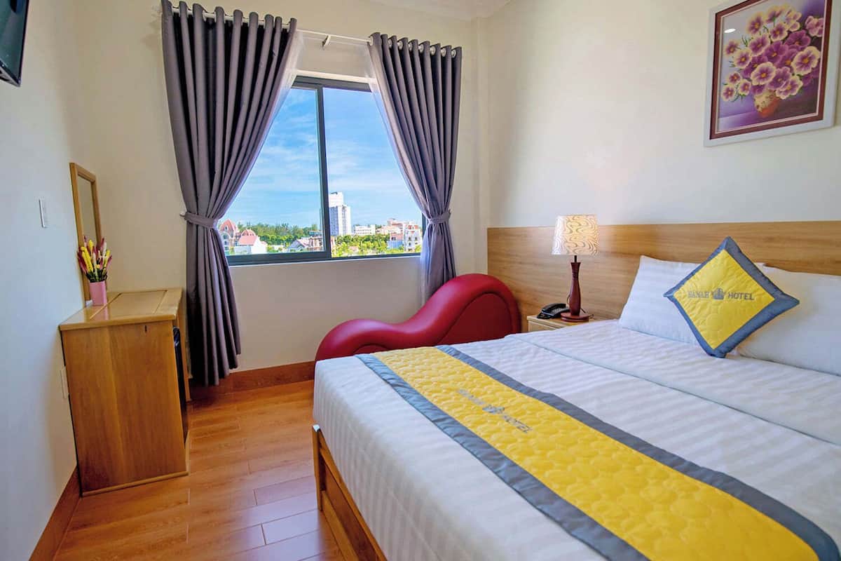 Hotellihuone, jossa on sänky, yöpöytä, punainen tuoli, puinen työpöytä, lamppu ja ikkuna, jossa on verhot.