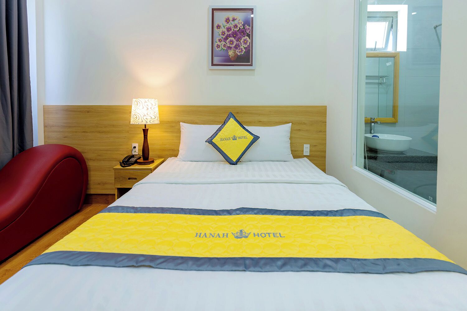 Une chambre d’hôtel avec un lit, une table de chevet avec une lampe, un fauteuil rouge et une salle de bain visible à travers une porte en verre.