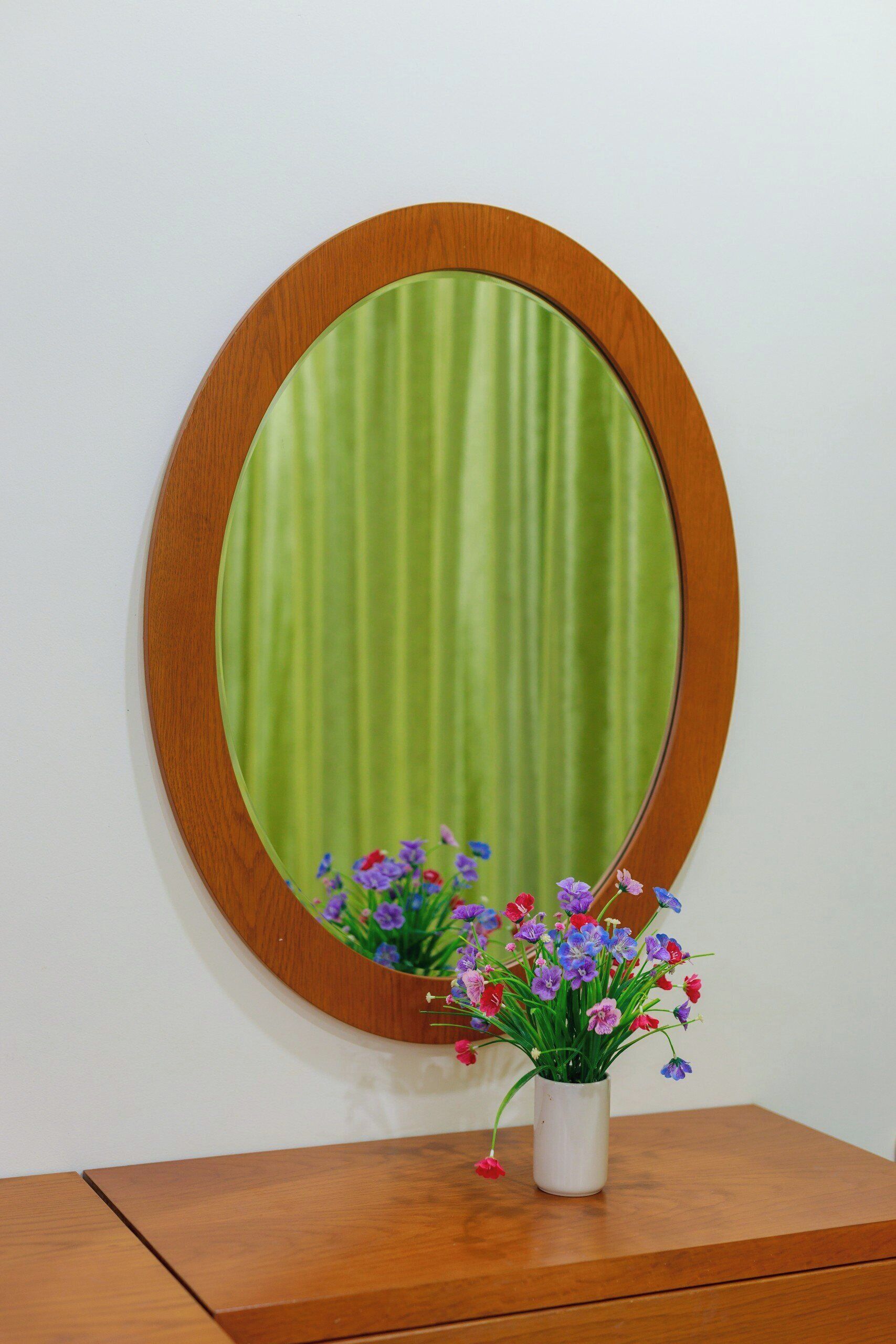 A wooden-framed oval mirror with a green curtain, reflecting colorful flowers in a vase on a wooden dresser.