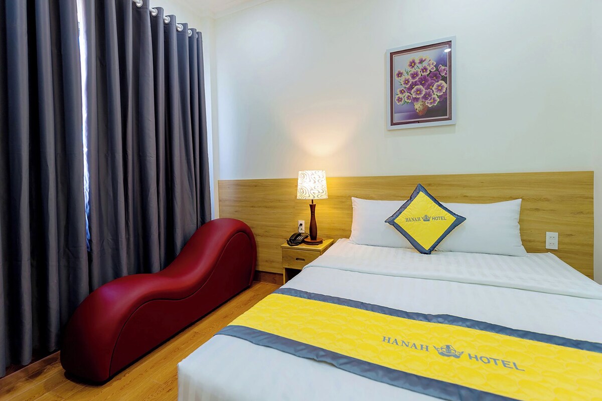 Habitación de hotel con cama, mesita de noche con lámpara, una silla roja y un cuadro en la pared.