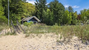 Une maison de plage avec un escalier menant à une terrasse en bois, entourée de sable et d’une végétation clairsemée.