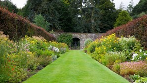 A garden pathway leading to a stone archway surrounded by diverse flowering plants and trees.