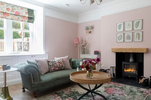 A cozy living room with a green sofa, a round wicker table with flowers, a fireplace, and framed botanical prints on the wall.