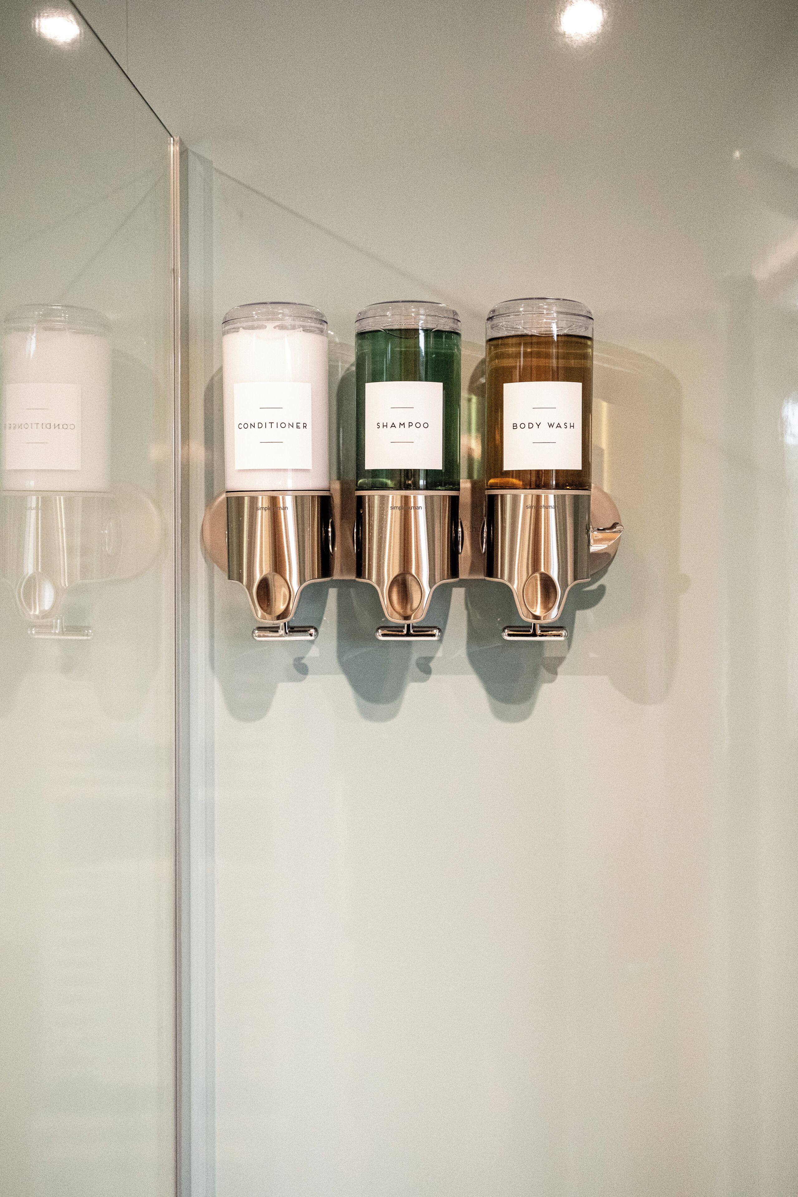 A wall-mounted dispenser with three containers labeled conditioner, shampoo, and body wash.