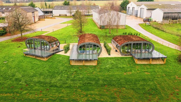 Un edificio moderno con tres grandes estructuras de vidrio curvas sobre un campo cubierto de césped, rodeado de construcciones agrícolas.
