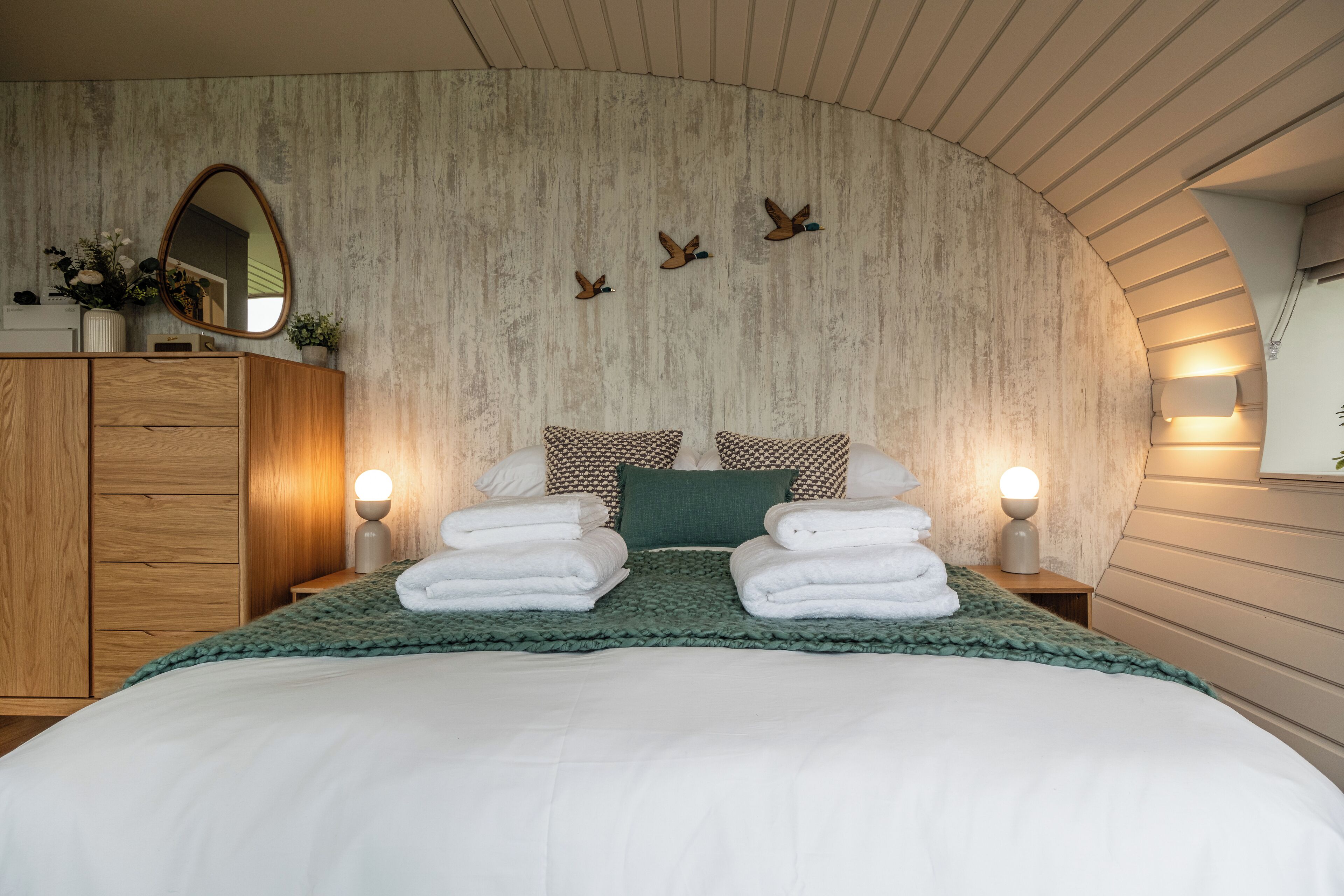A neatly made bed with white linens and a green throw blanket, flanked by two bedside lamps and a mirror on the wall.