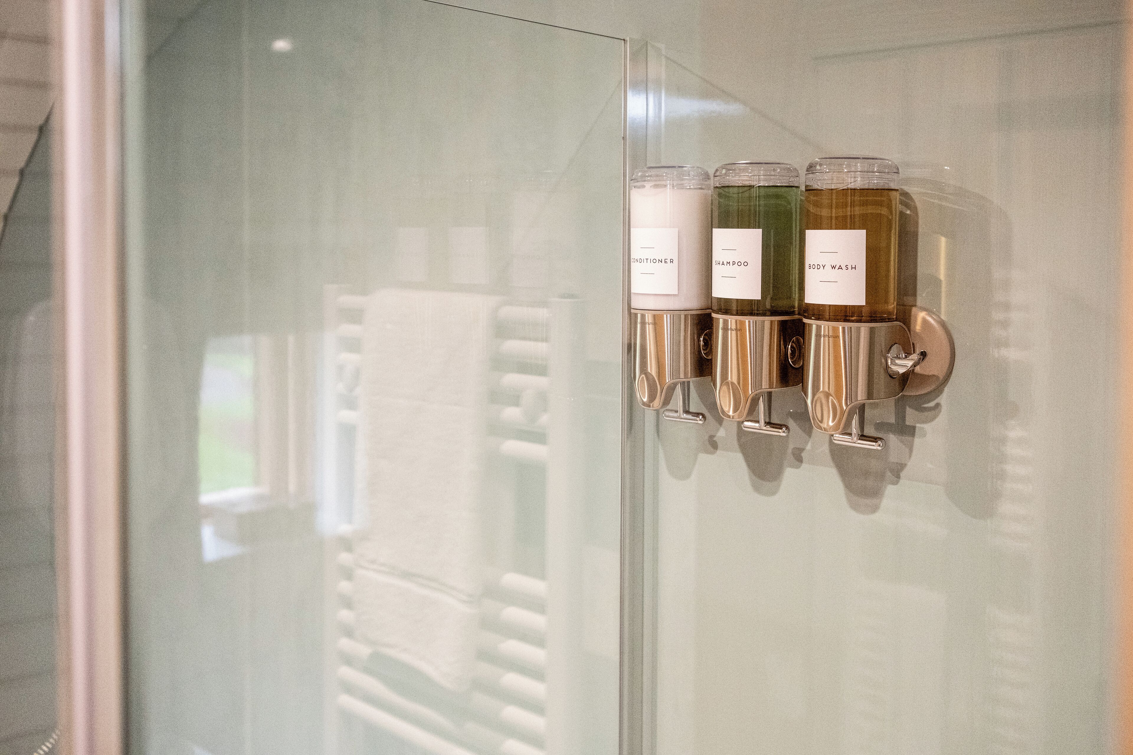 A bathroom with a glass door and three bottles of toiletries labeled "Shampoo," "Body Wash," and "Conditioner."