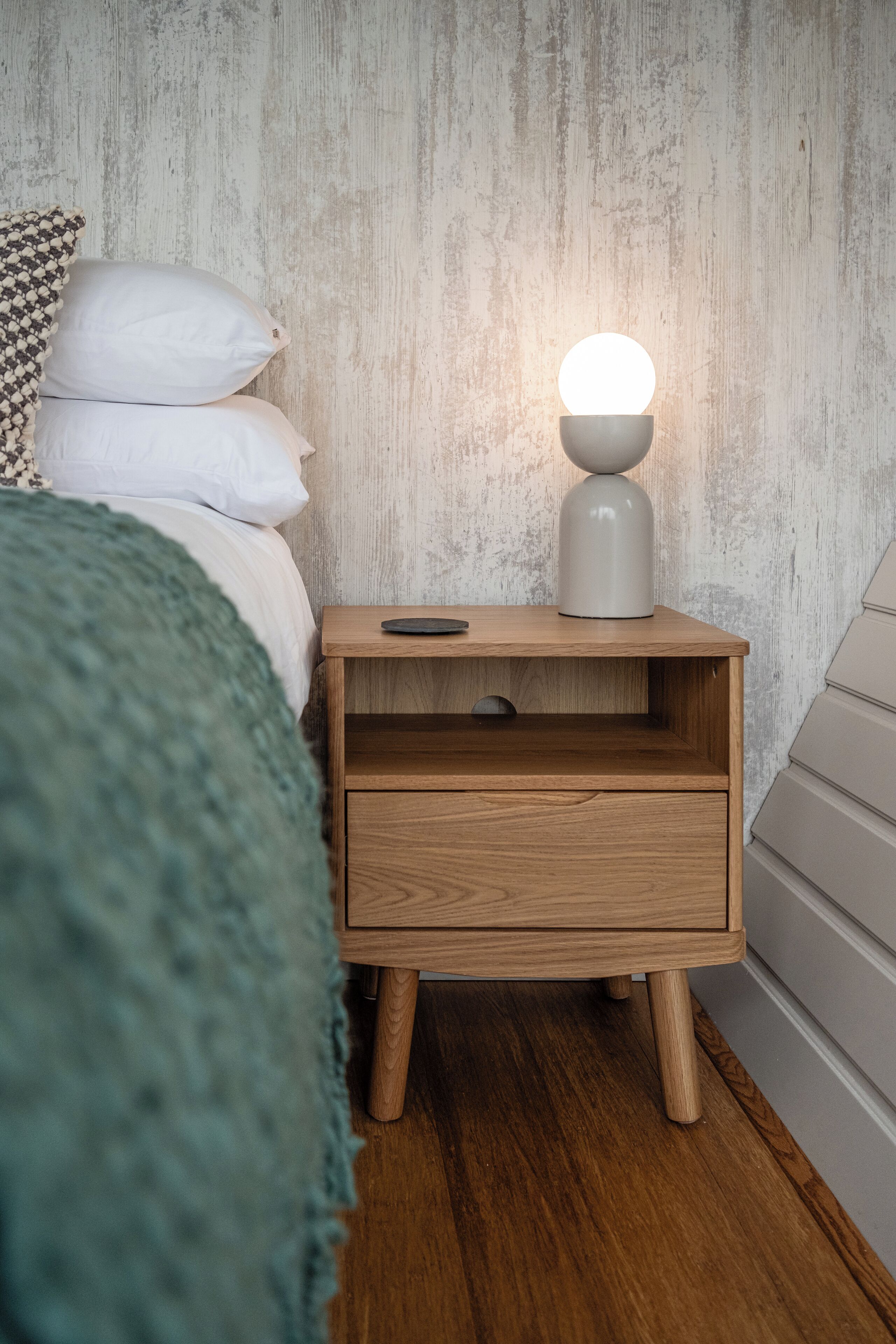 A wooden nightstand with a lamp, a bed with white pillows, and a textured wall.