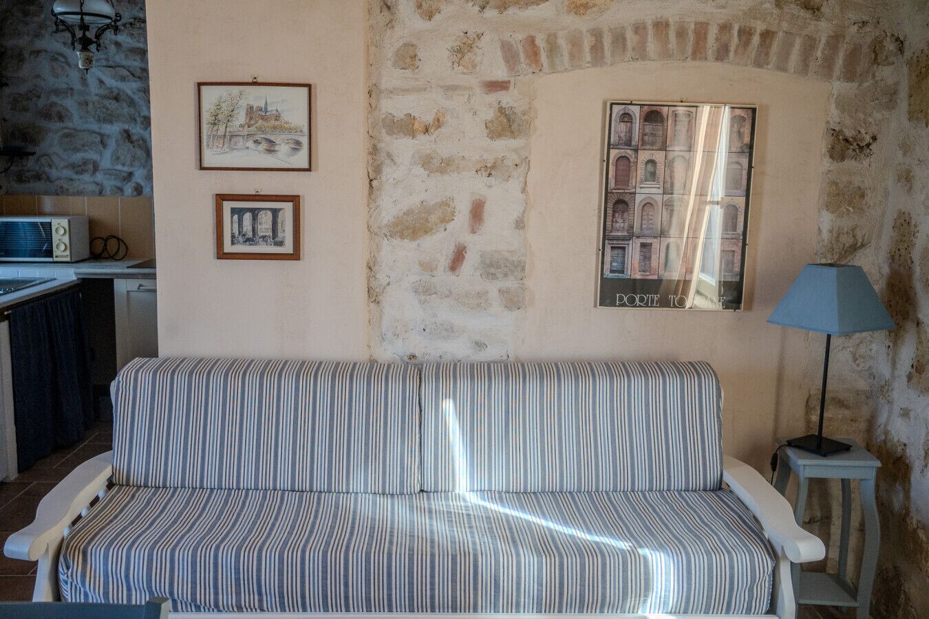 Ein Zimmer mit einem gestreiften Sofa, einem kleinen Tisch mit Lampe, einer Küchenzeile und zwei gerahmten Bildern an der Wand.